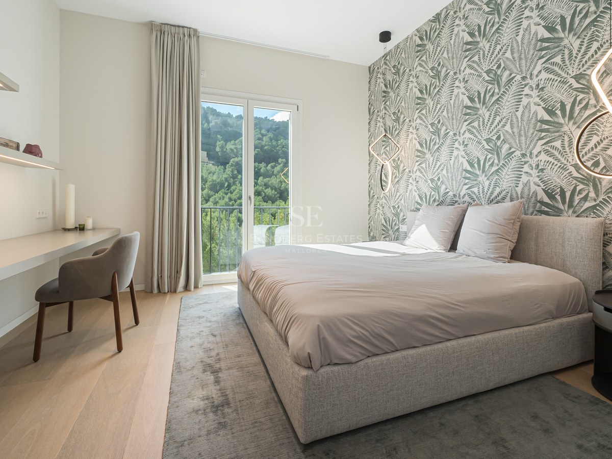 Dormitorio contemporáneo con papel pintado botánico, cama tapizada, escritorio integrado y balcón con vistas a colinas.