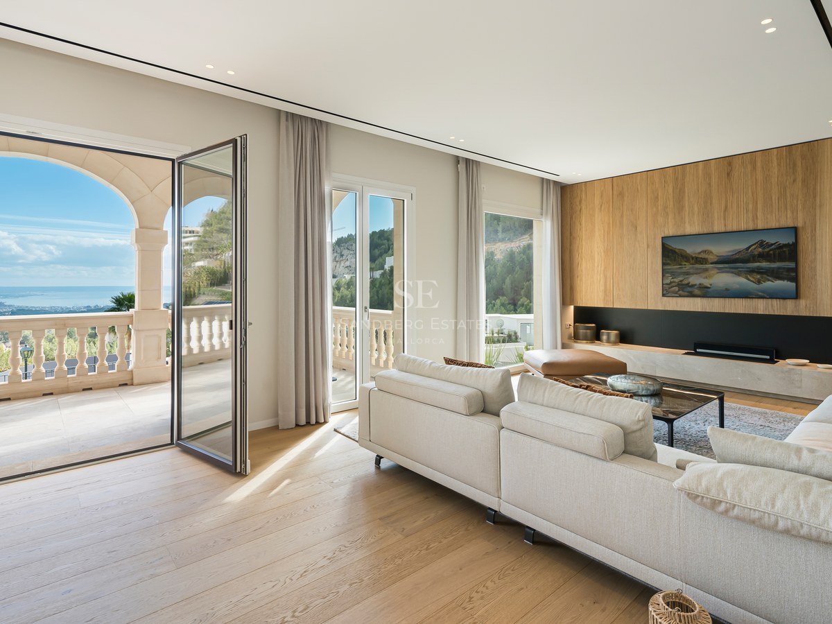 Salón espacioso con suelos de roble, sofá crema y grandes puertas de cristal que dan a una terraza con vistas al mar.
