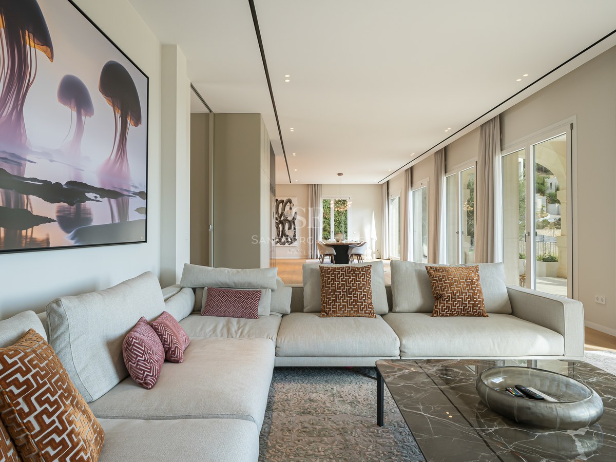 Salón moderno con sofá en L color crema, mesa de mármol y grandes ventanales con vistas al exterior.