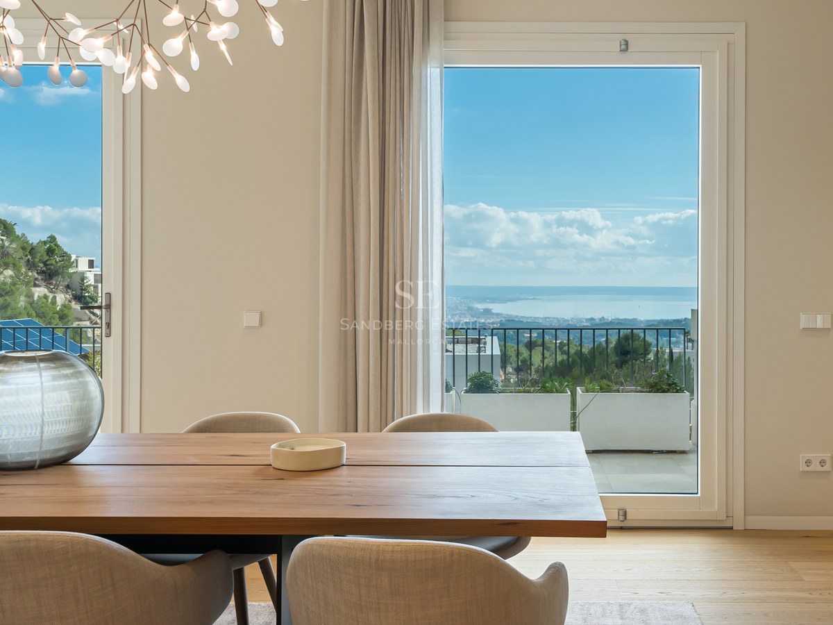 Comedor moderno con mesa de madera maciza y grandes ventanales con vistas a la costa mediterránea.