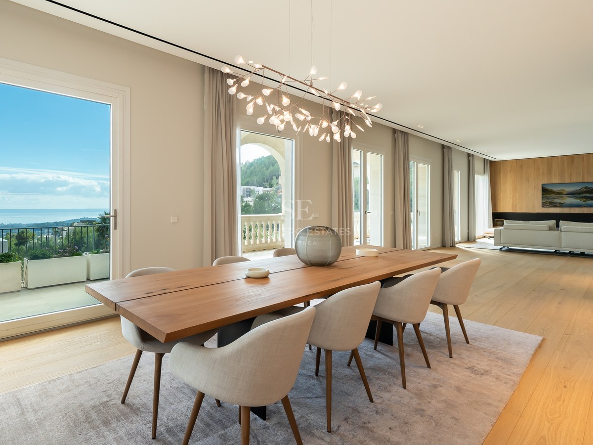 Elegante comedor con mesa de madera maciza, lámpara escultórica y grandes ventanales con vistas al mar y la costa.