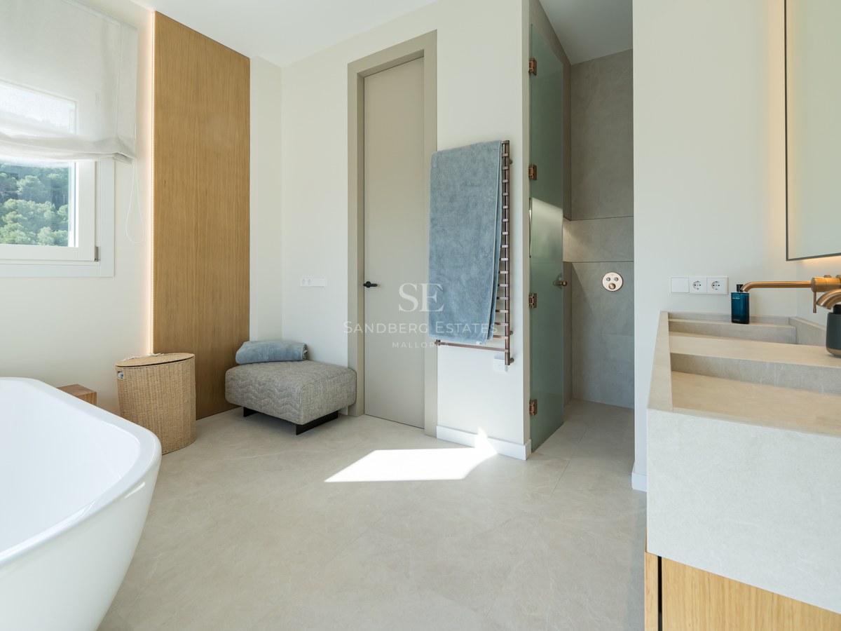 Baño moderno con bañera exenta, lavabo doble de piedra natural, grifería de bronce y vistas al bosque.