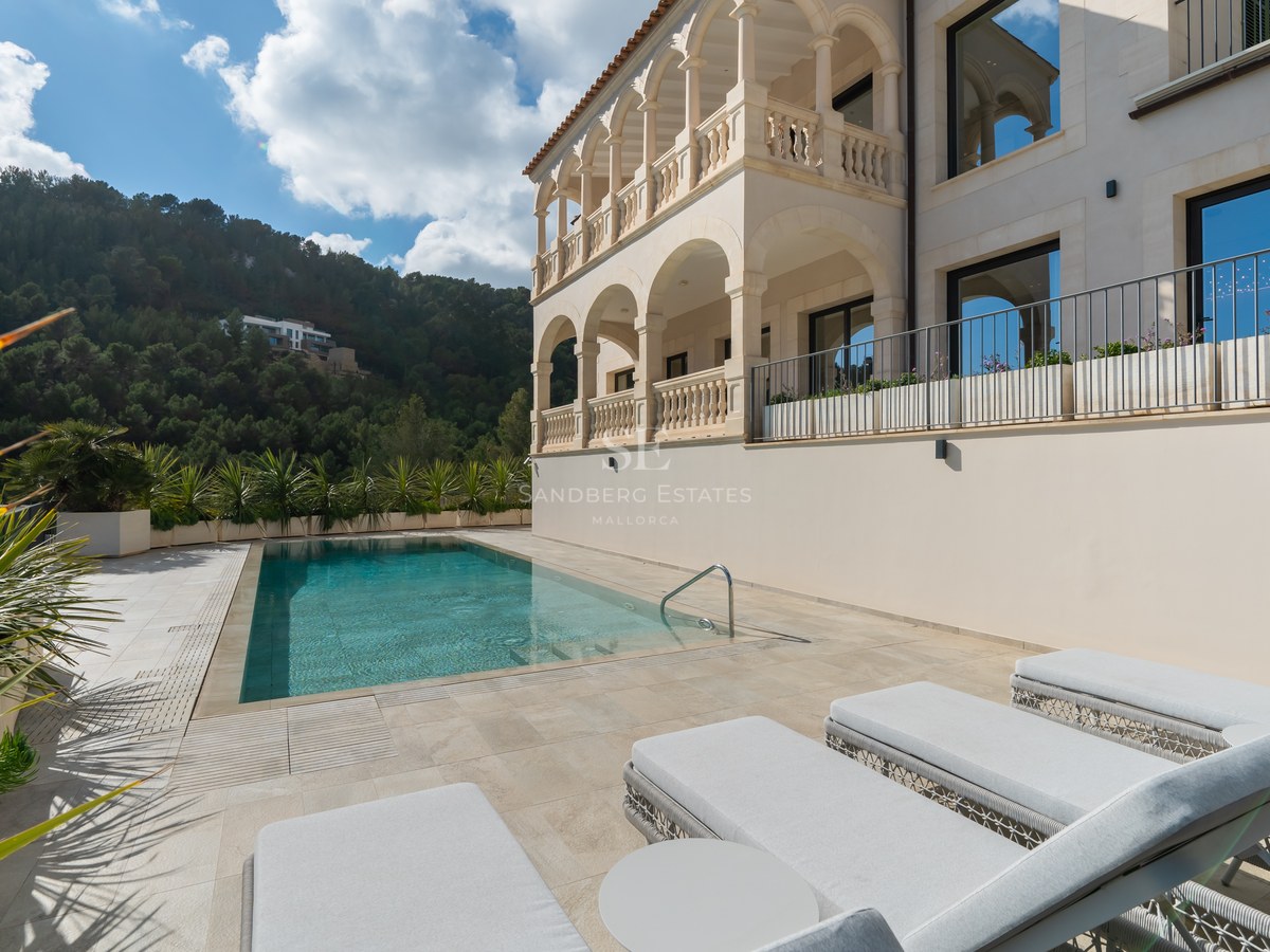 Villa de lujo con piscina rectangular, tumbonas, balcones de piedra con arcos y colinas verdes al fondo.