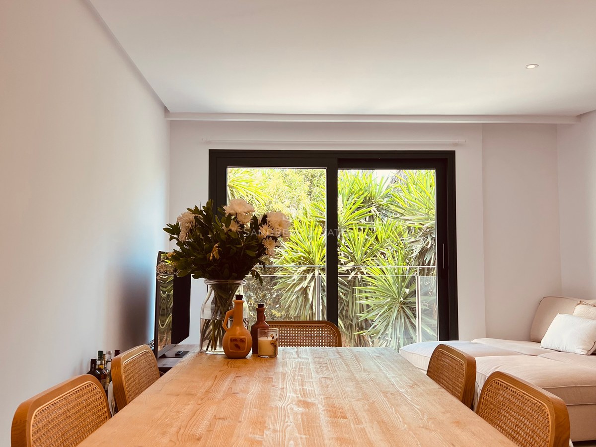 Light wood dining table with rattan chairs in front of a large window looking onto tropical greenery.
