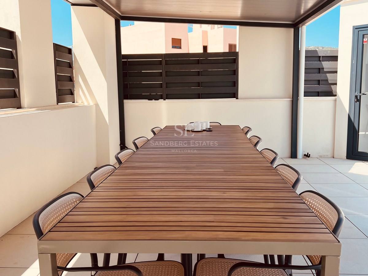 A long wooden outdoor dining table with ten chairs under a modern pergola on a sunlit terrace.