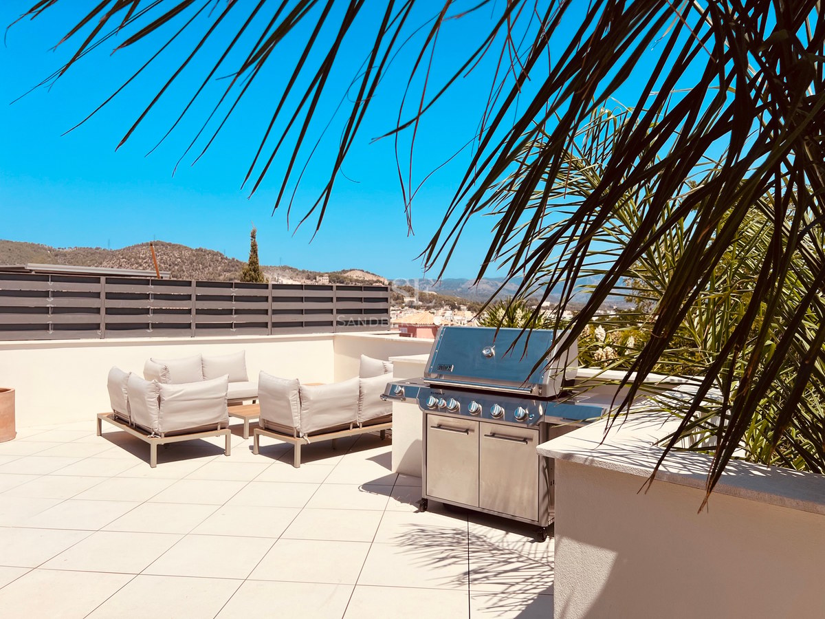 Sun-drenched rooftop terrace featuring a large stainless steel grill, modern lounge seating, and mountain backdrop.