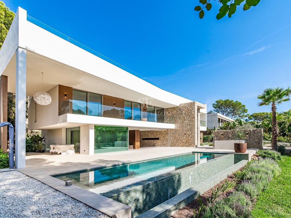 Modern two-story villa facade with infinity pool, natural stone walls, and large glass windows under a clear blue sky.