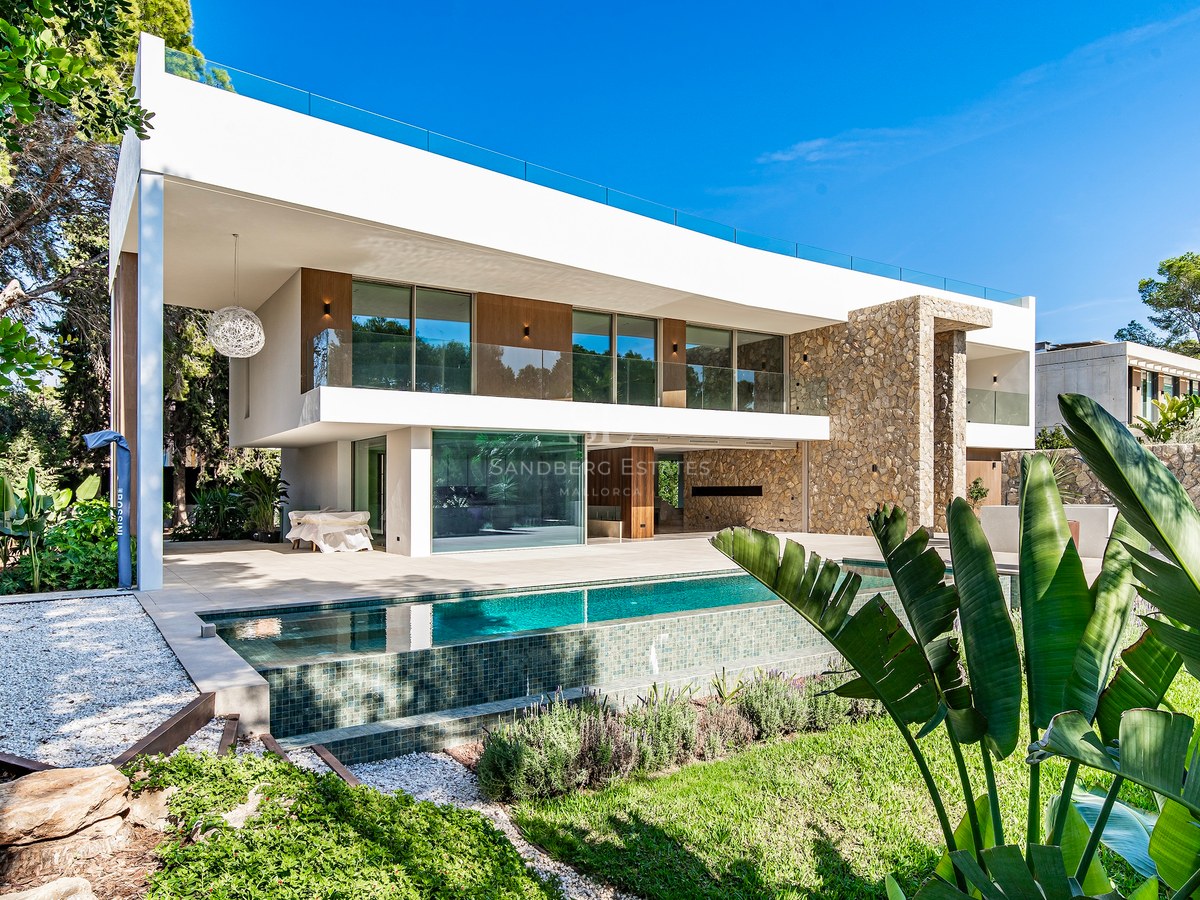Two-story white modern villa with natural stone walls, a large swimming pool, and glass railings under a clear blue sky.