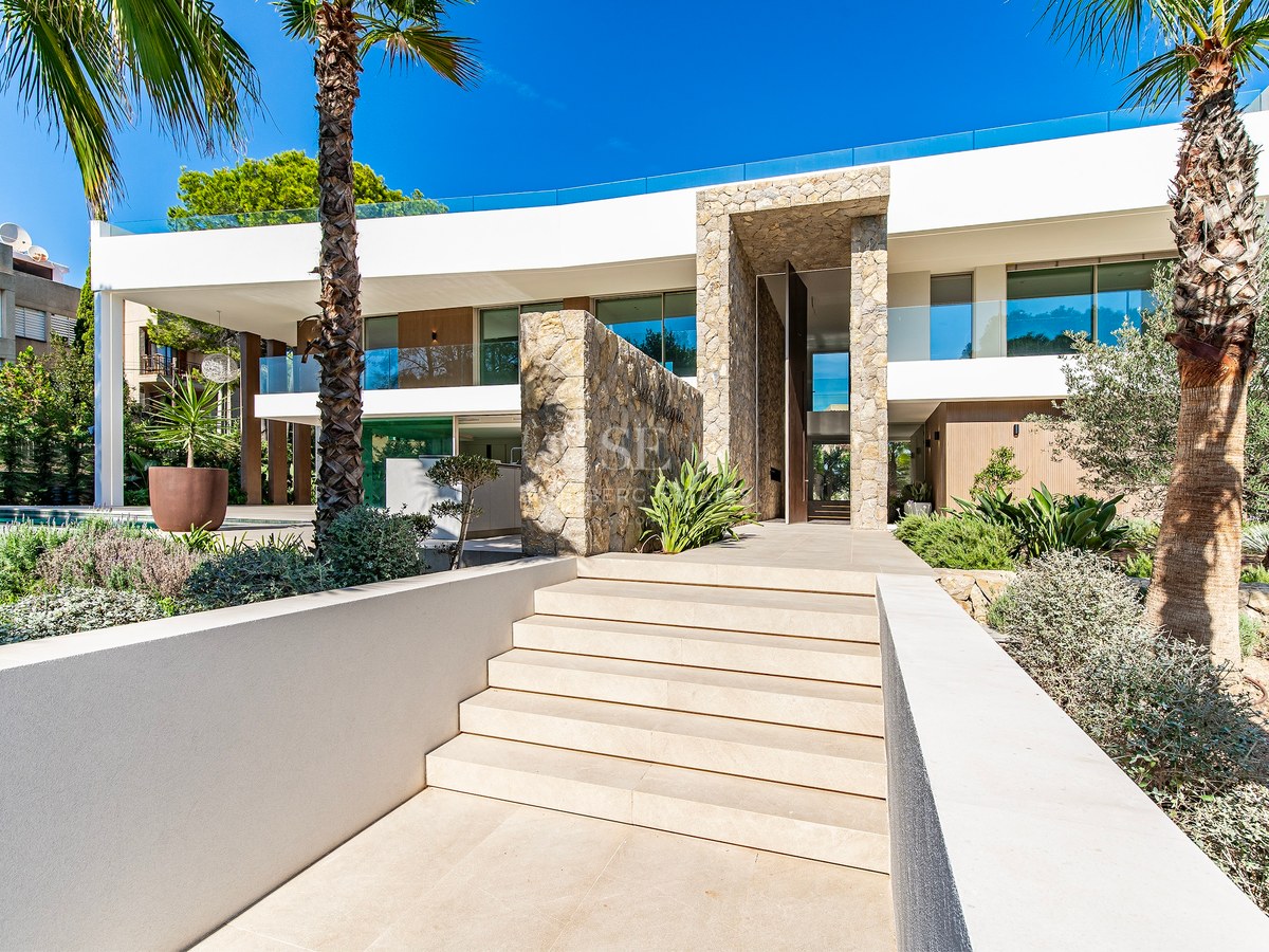 Modern white villa with natural stone feature walls, stone steps, and palm trees under a bright blue sky.