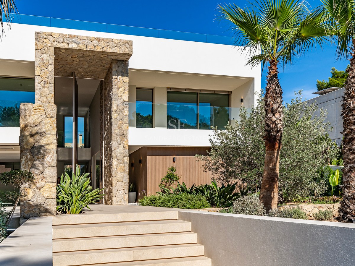 Modern villa entrance featuring wide stone steps, a high stone-clad portal, palm trees, and clear blue sky.