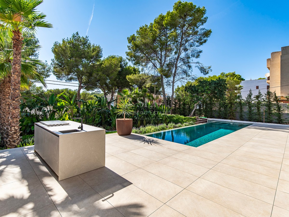 Stone terrace featuring a minimalist outdoor kitchen island next to a turquoise swimming pool and tropical gardens.