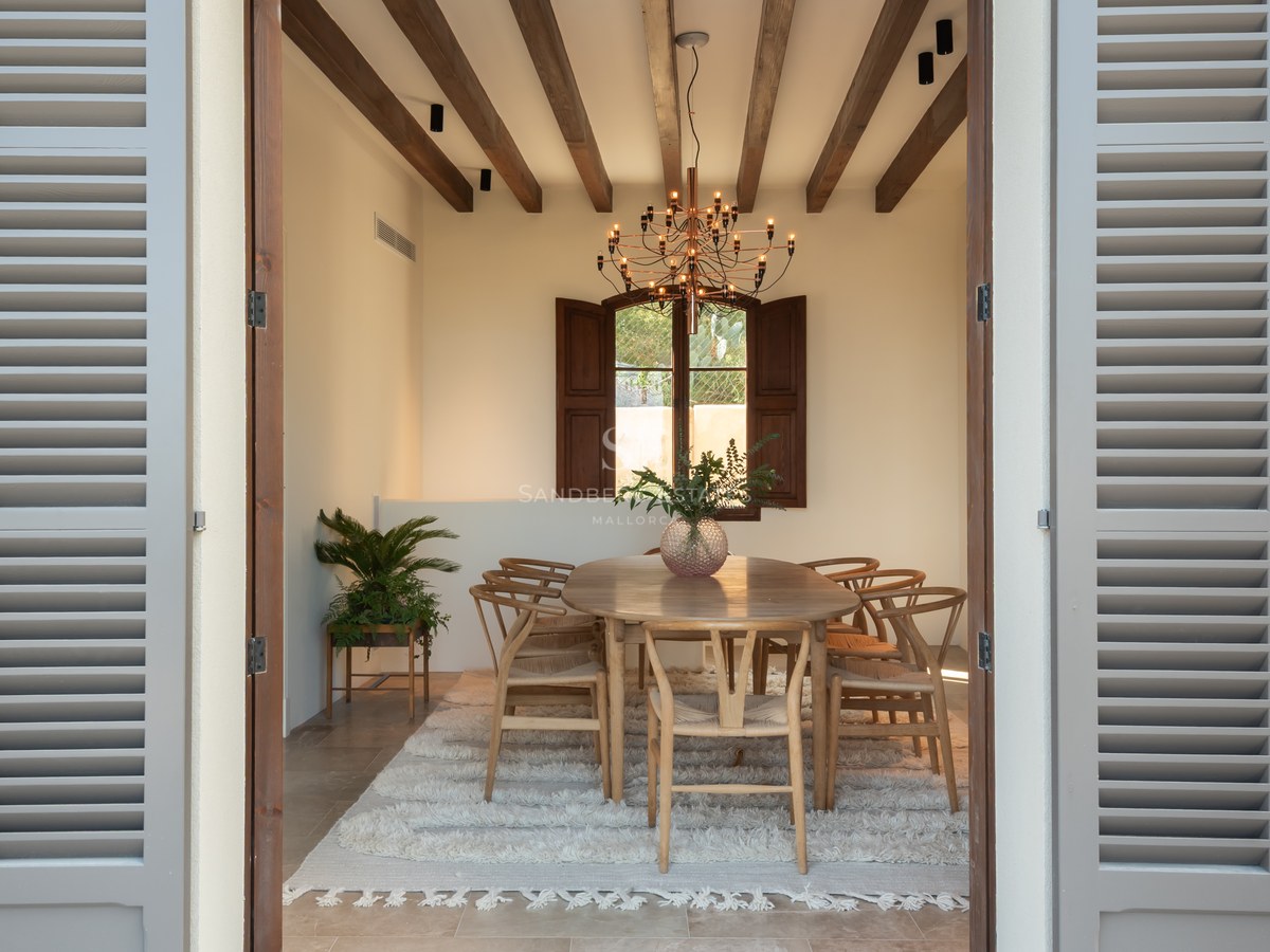 Salle à manger avec poutres apparentes, table en chêne, chaises Wishbone et tapis blanc encadrée par des volets gris.