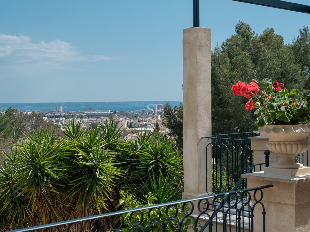 Vue depuis une terrasse sur une ville méditerranéenne et la mer, avec fleurs rouges et balustrade en fer forgé.