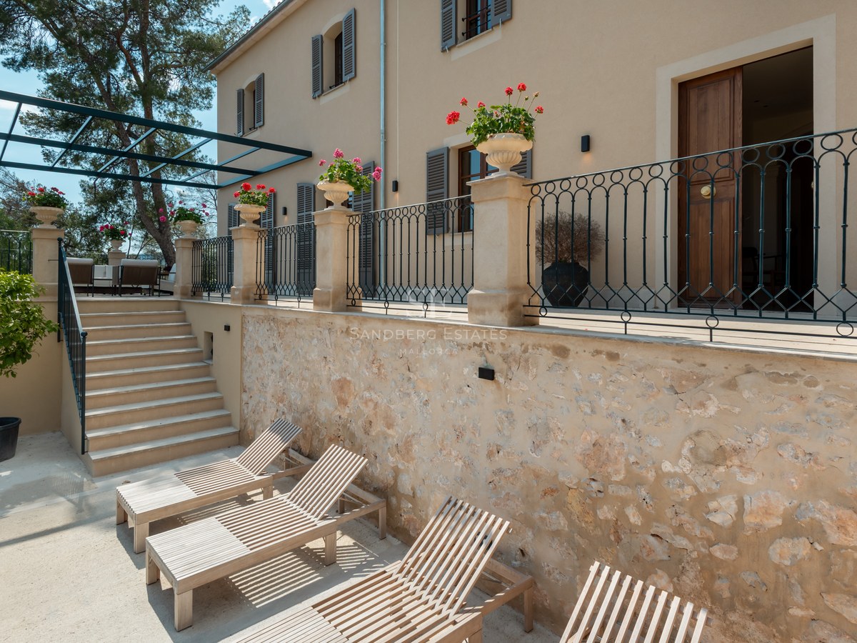 Terrasse en pierre à plusieurs niveaux avec chaises longues en bois, balustrades en fer forgé et façade de villa.