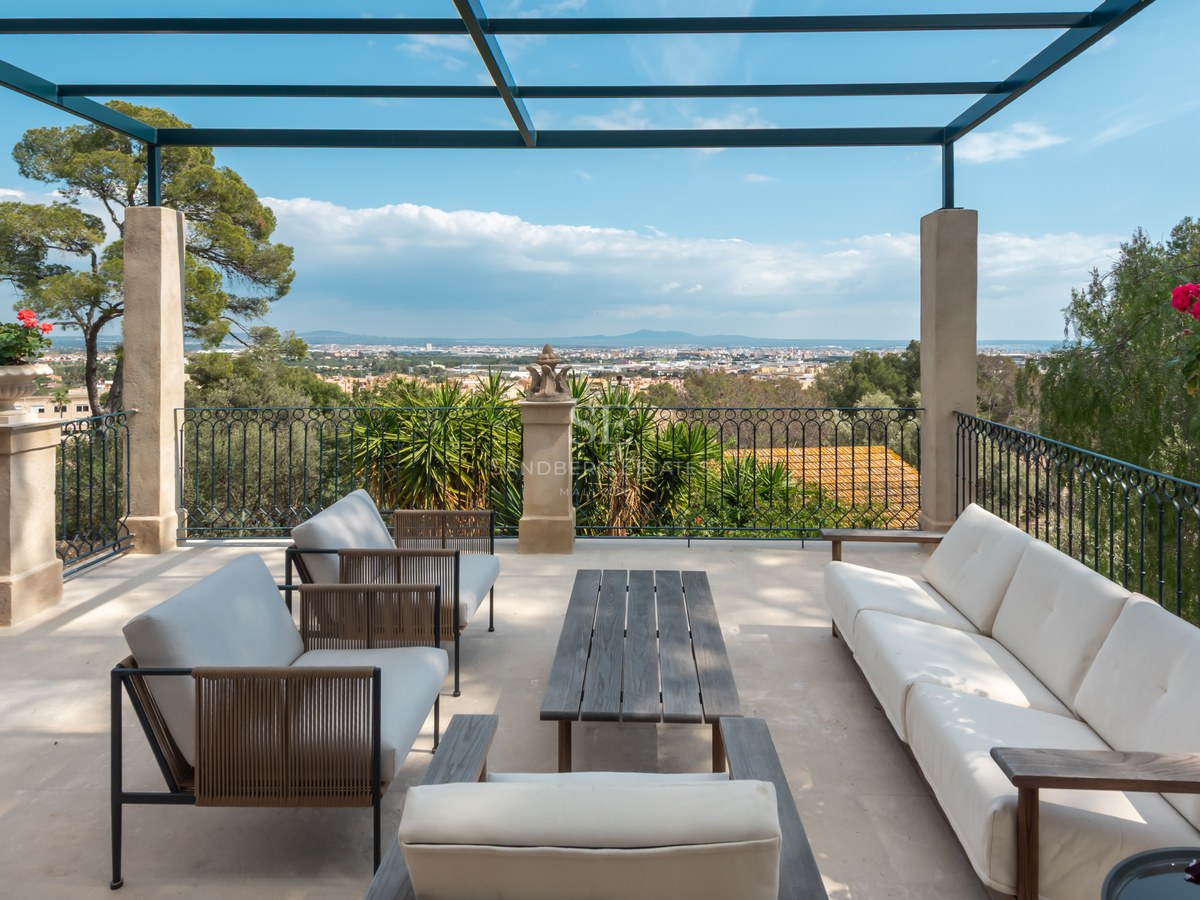 Terrasse de luxe avec canapés blancs, piliers en pierre et balustrade en fer forgé surplombant une vallée et des montagnes.