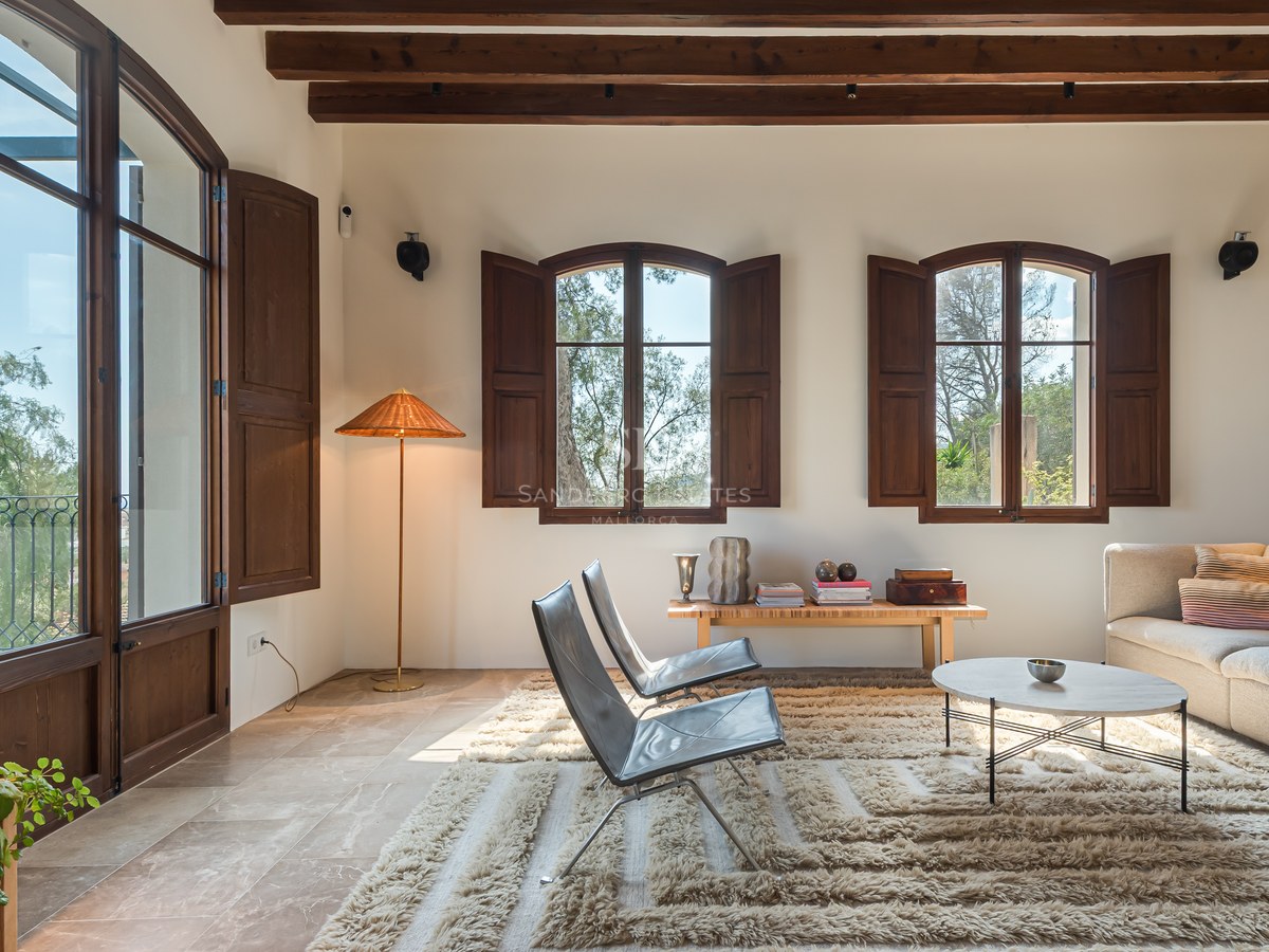 Salon lumineux avec sol en pierre, poutres en bois sombre, fenêtres cintrées avec volets et mobilier moderne.