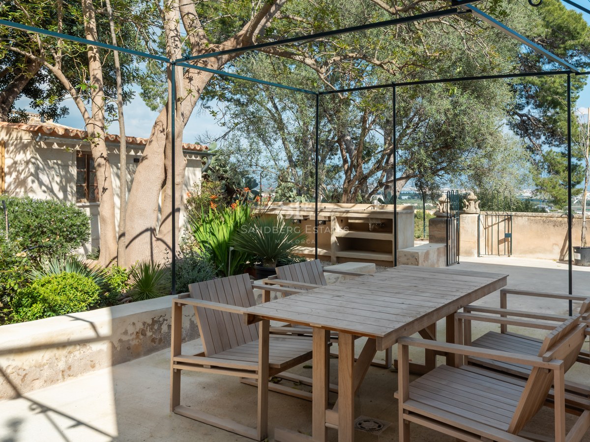Espace dînatoire en bois sur terrasse en pierre avec pergola métallique, cuisine d'été et arbres matures.