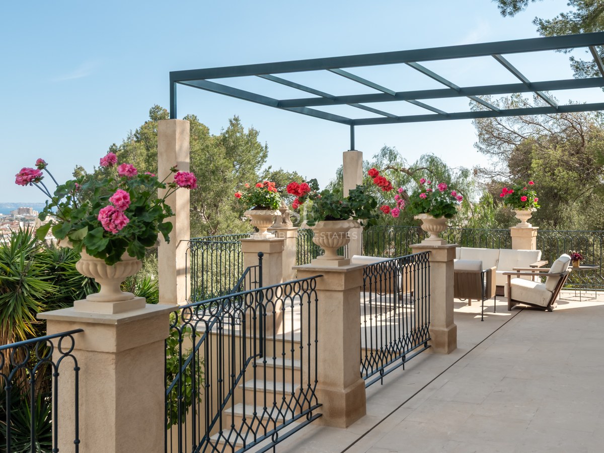 Terrasse en pierre avec garde-corps en fer, pergola métallique et fleurs surplombant la ville et la mer.
