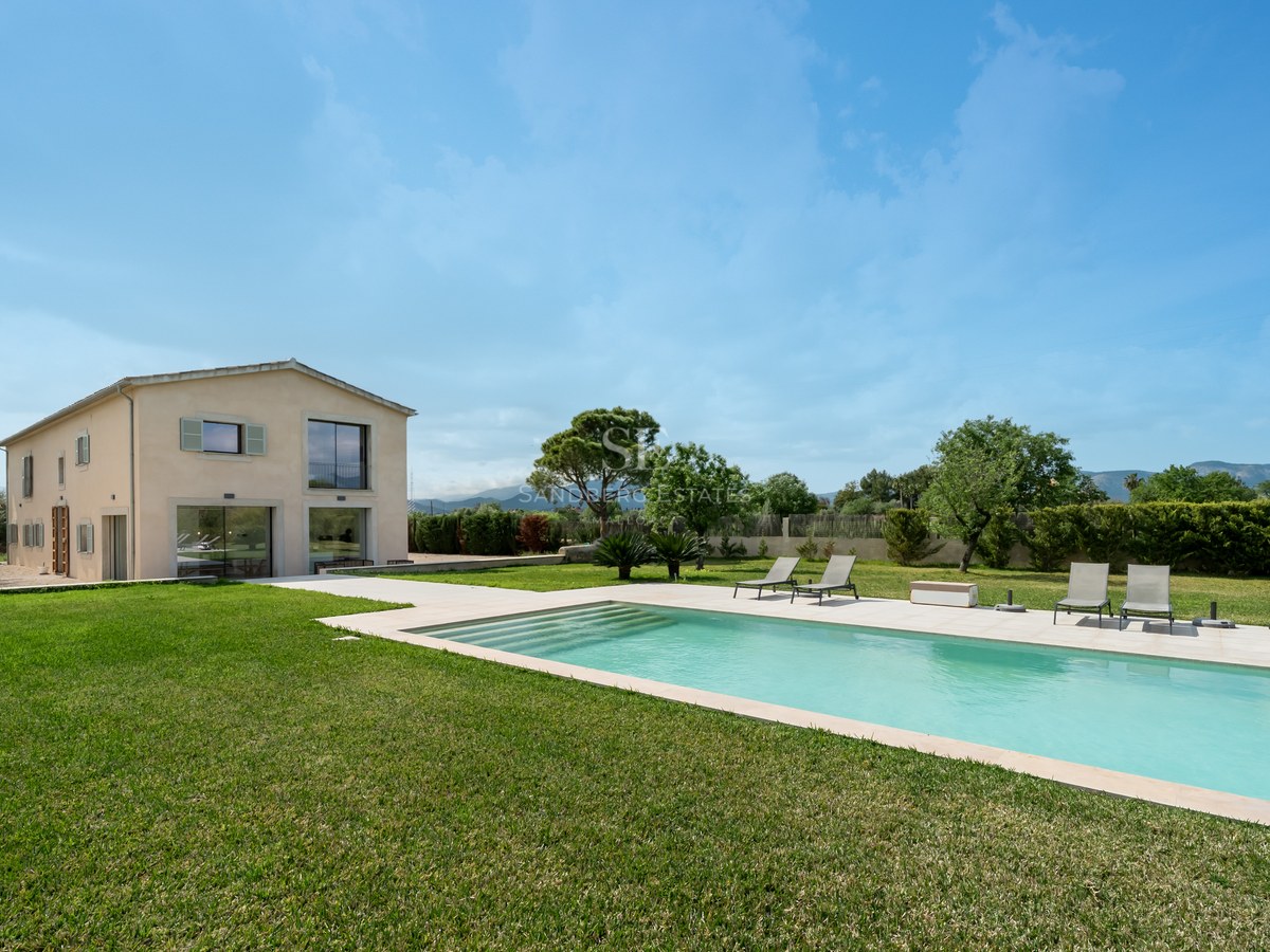 Rektangulär pool med solstolar på en grön gräsmatta framför en modern beige villa under en klarblå himmel.