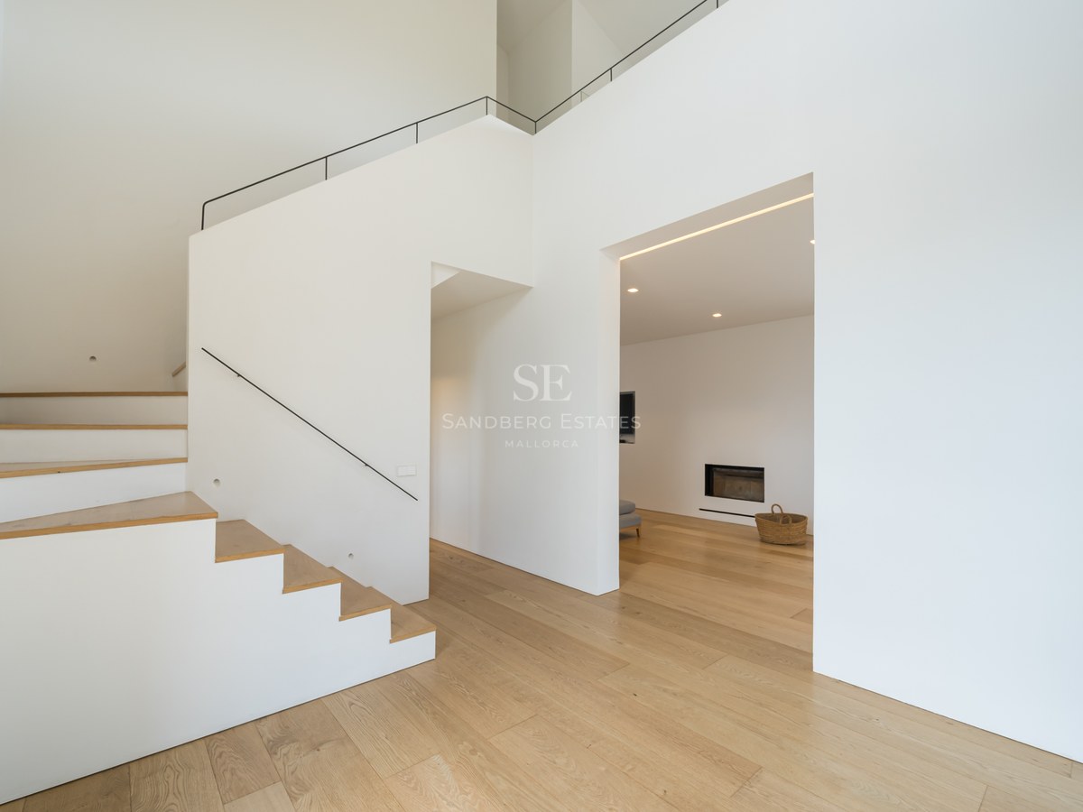 Modern minimalistisk hall med vita väggar, ljust ekparkettgolv och stilren vit trappa med svart räcke.