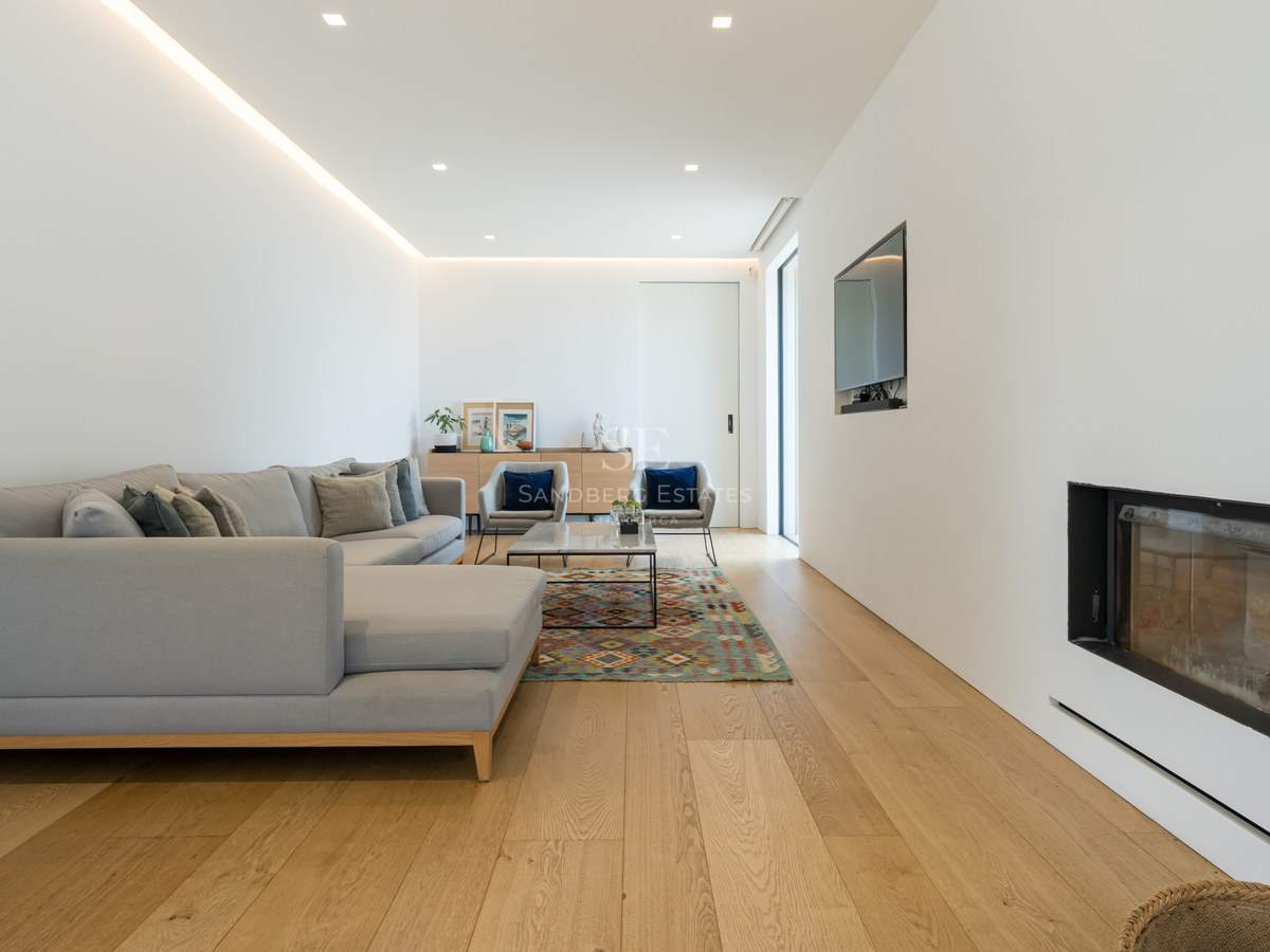 Ljust vardagsrum med ekparkett, grå L-sofa, inbyggd öppen spis och infälld LED-belysning.