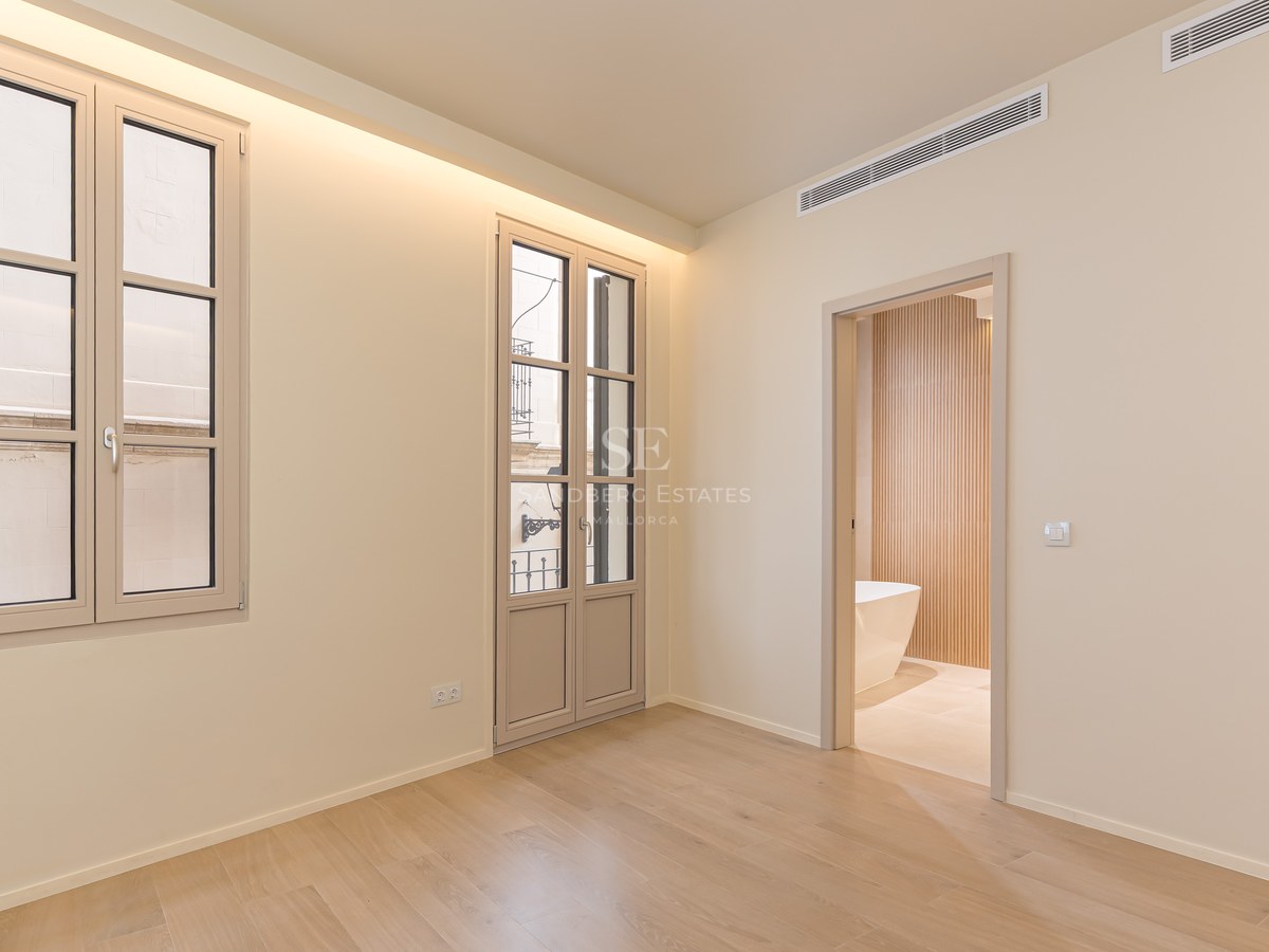 Dormitorio minimalista con suelos de madera clara, ventanales altos y vista a un baño con bañera exenta y listones de madera.