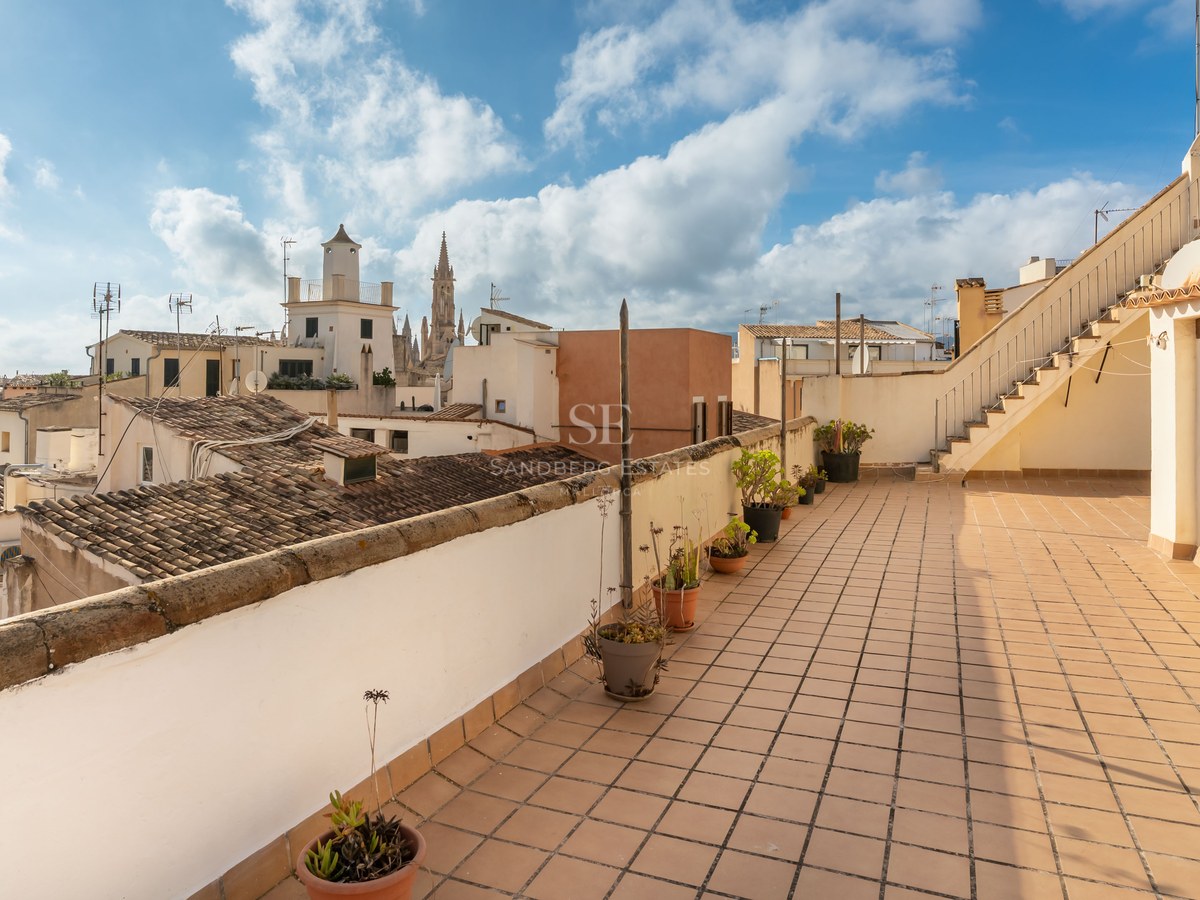 Terraza en azotea con baldosas de terracota con vistas a edificios históricos y un campanario gótico bajo un cielo azul.