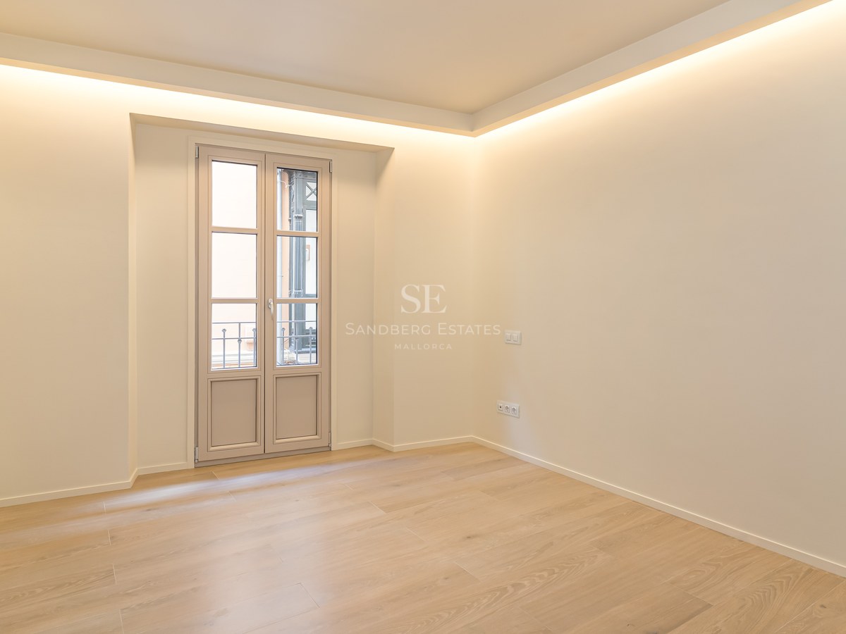 Habitación moderna vacía con suelos de roble claro, paredes crema, iluminación LED perimetral y balcón francés.