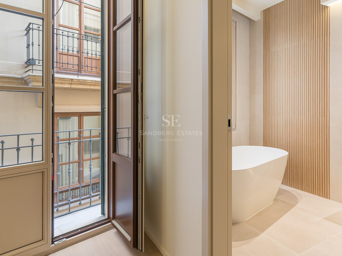 Baño moderno con bañera blanca exenta, lavabo de piedra, pared de listones de madera y balcón hacia patio exterior.