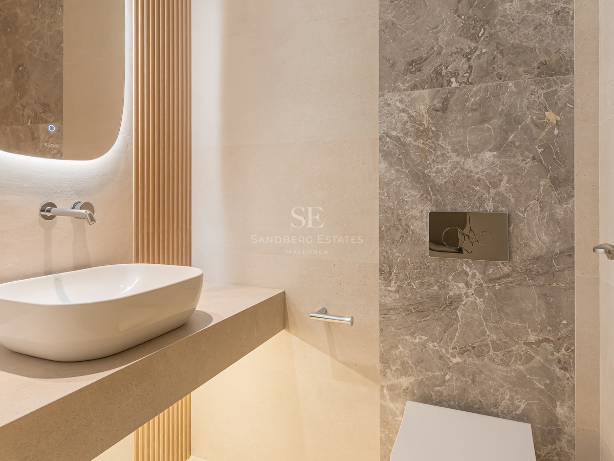 Aseo moderno con lavabo sobre encimera, espejo retroiluminado, listones de madera y pared de mármol gris.