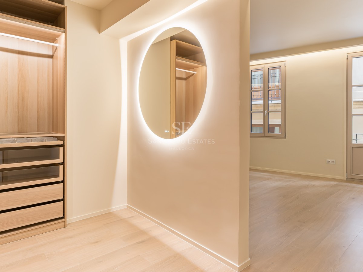 Vestidor de lujo con armarios de roble claro, espejo circular retroiluminado y cajones con frente de cristal.