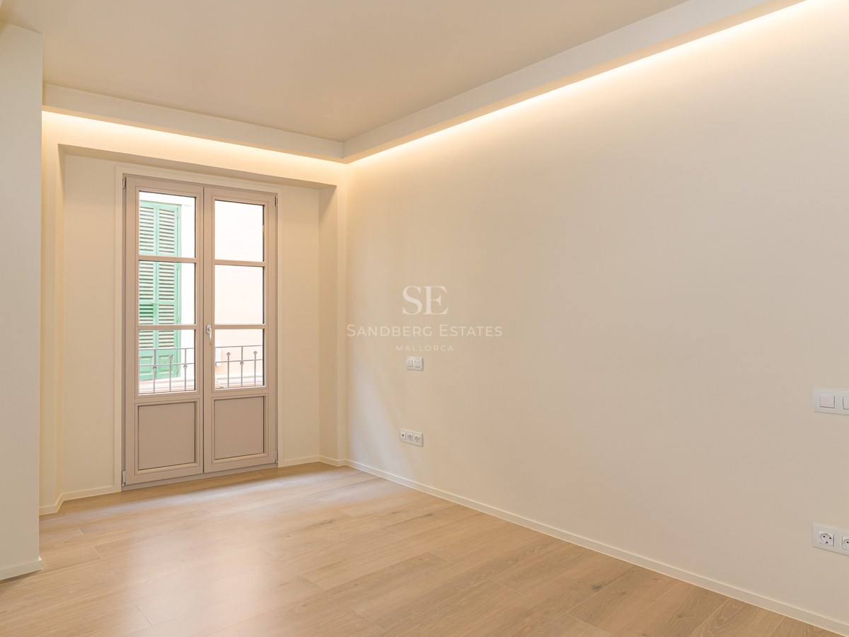 Dormitorio vacío con suelo de roble claro, paredes crema, iluminación indirecta y puerta hacia balcón francés.