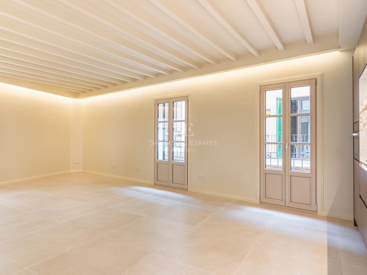 Salón vacío y luminoso con suelo de gres crema, techo de vigas vistas, tres puertas balconeras y cocina moderna lateral.