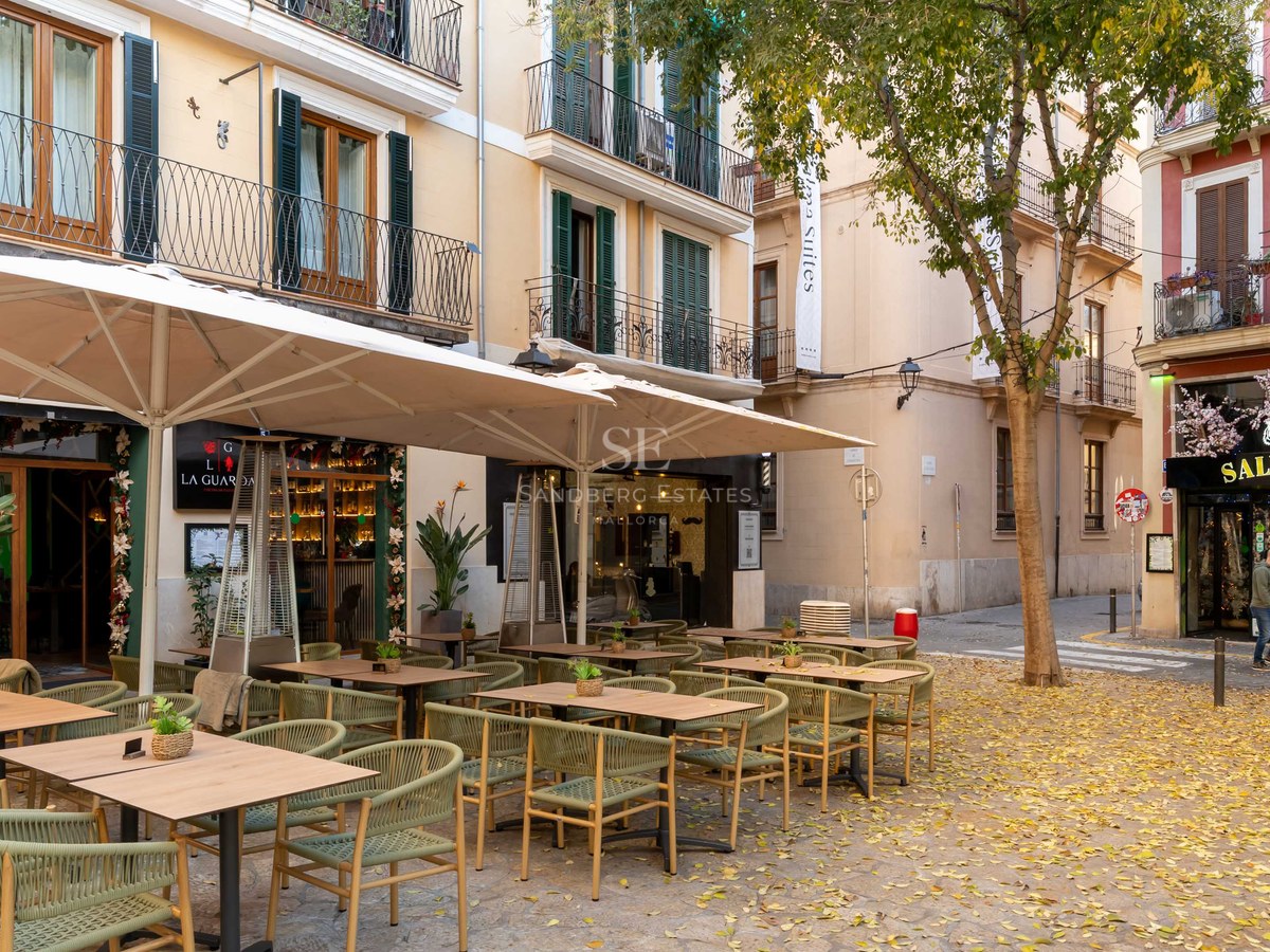 Terraza de restaurante con sillas verde salvia bajo sombrillas en una calle histórica mediterránea con hojas de otoño.