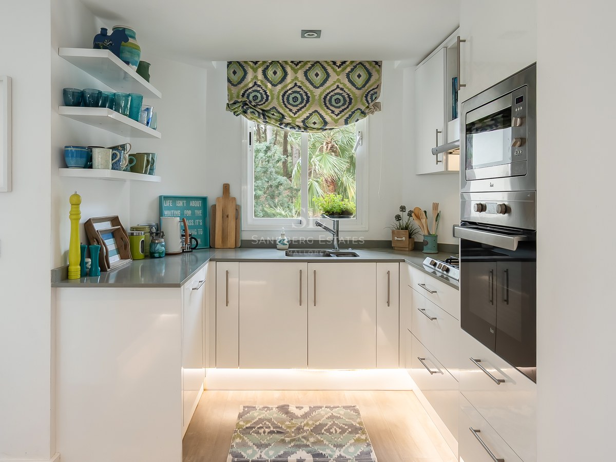 Bright U-shaped kitchen with white cabinets, stainless steel appliances, and a window overlooking lush green trees.