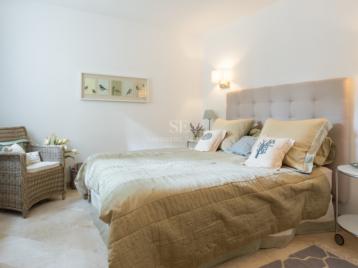 A bright bedroom featuring a tufted headboard, sage green bedding, marble floors, and a wicker armchair.