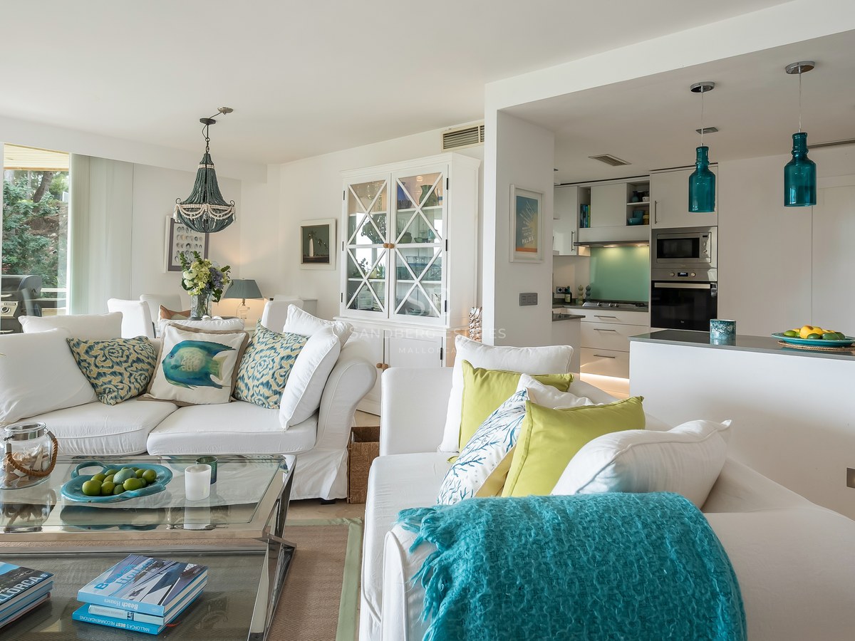 A sun-drenched living room with white sofas, turquoise accents, and a modern kitchen with blue pendant lights.