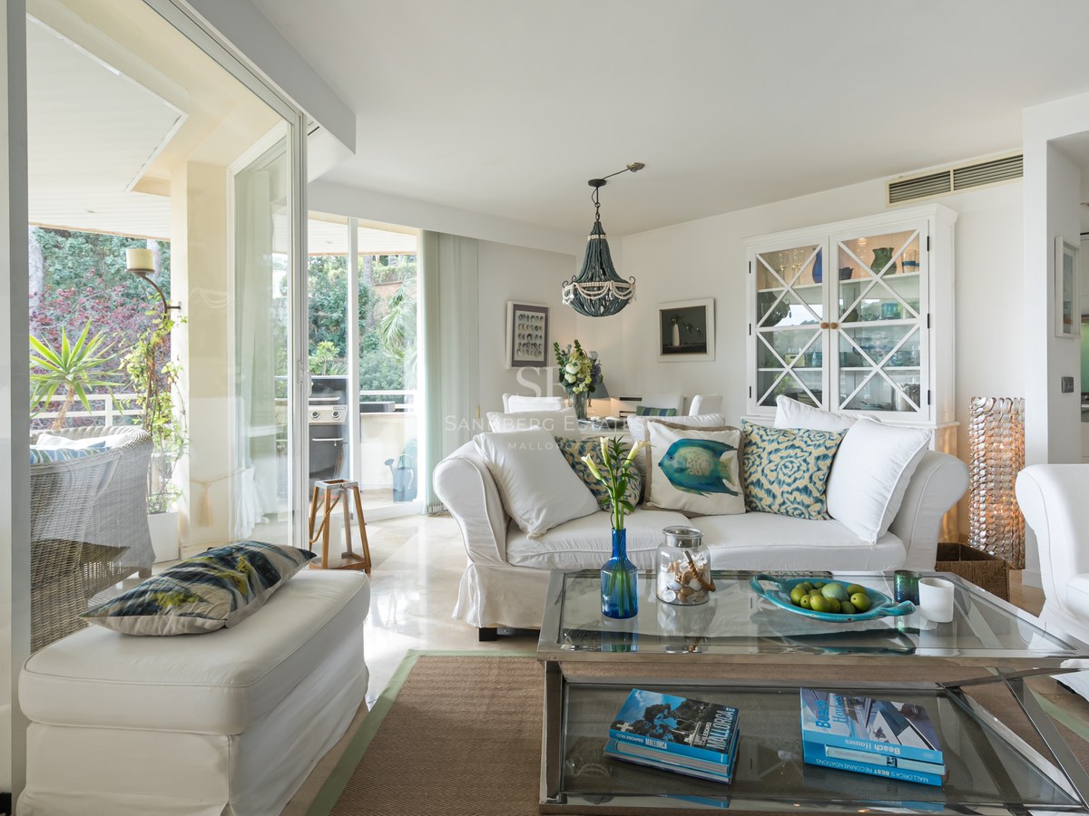 Open-plan living room featuring white sofas, a glass coffee table, and large sliding glass doors leading to a terrace.