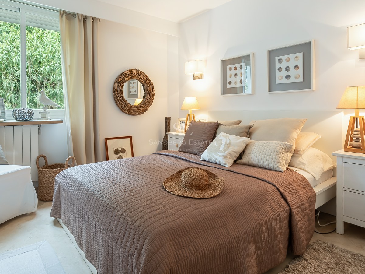 Bright bedroom with brown quilted bed, white nightstands, woven wall mirror, and garden views through a large window.
