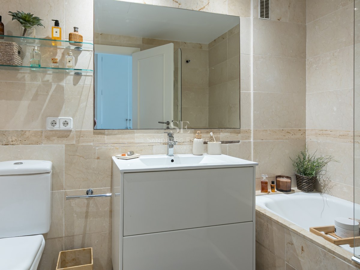 Bright bathroom featuring cream marble wall tiles, a white vanity unit, large mirror, and a bathtub with glass partition.
