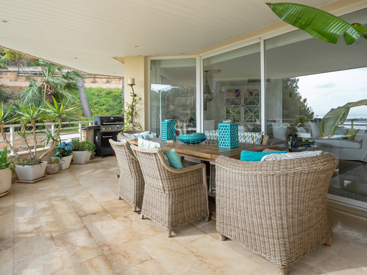 A shaded terrace featuring a wooden dining table, wicker armchairs with turquoise cushions, potted plants, and a grill.