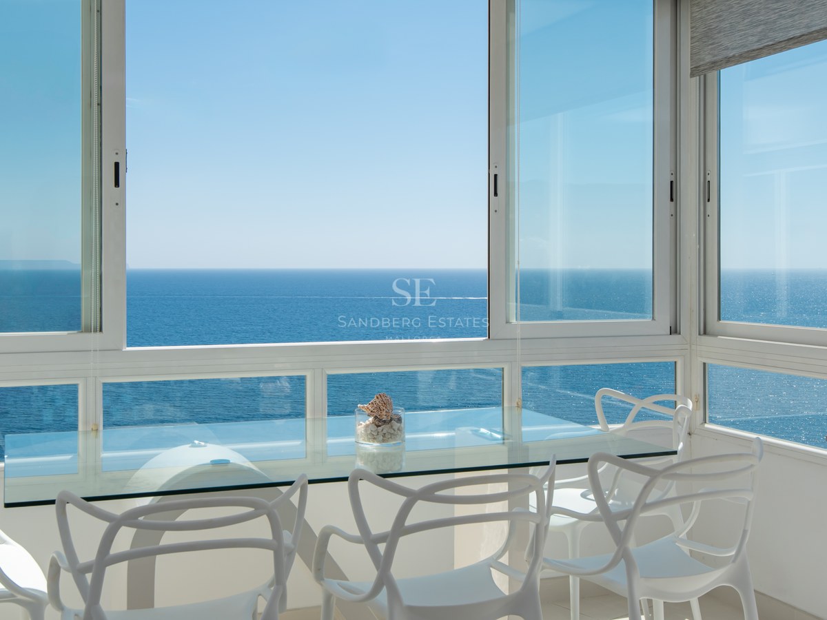 Modern dining area with white designer chairs and glass table overlooking the calm blue Mediterranean sea.