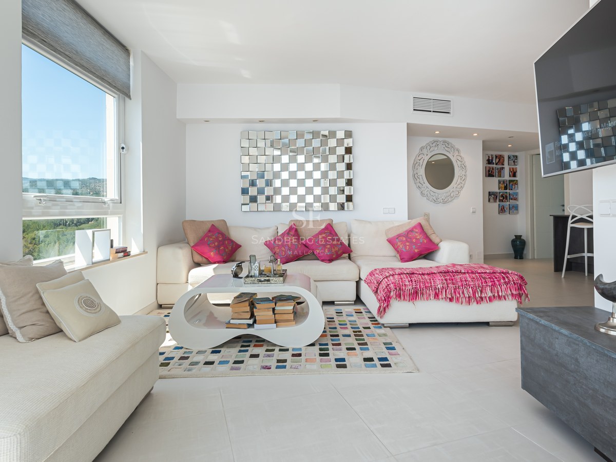 Bright living room with white L-shaped sofa, colorful rug, large window, and wall-mounted television.