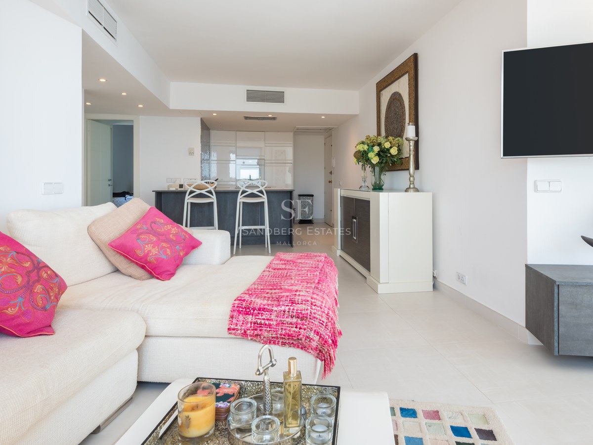 Bright living room with white L-shaped sofa, pink cushions, wall-mounted TV, and an open-plan kitchen in the background.