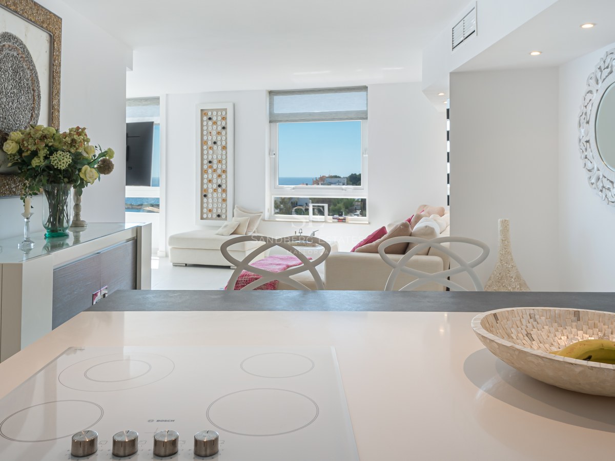 Modern white kitchen island looking into a bright living room with a sea view window and contemporary furniture.
