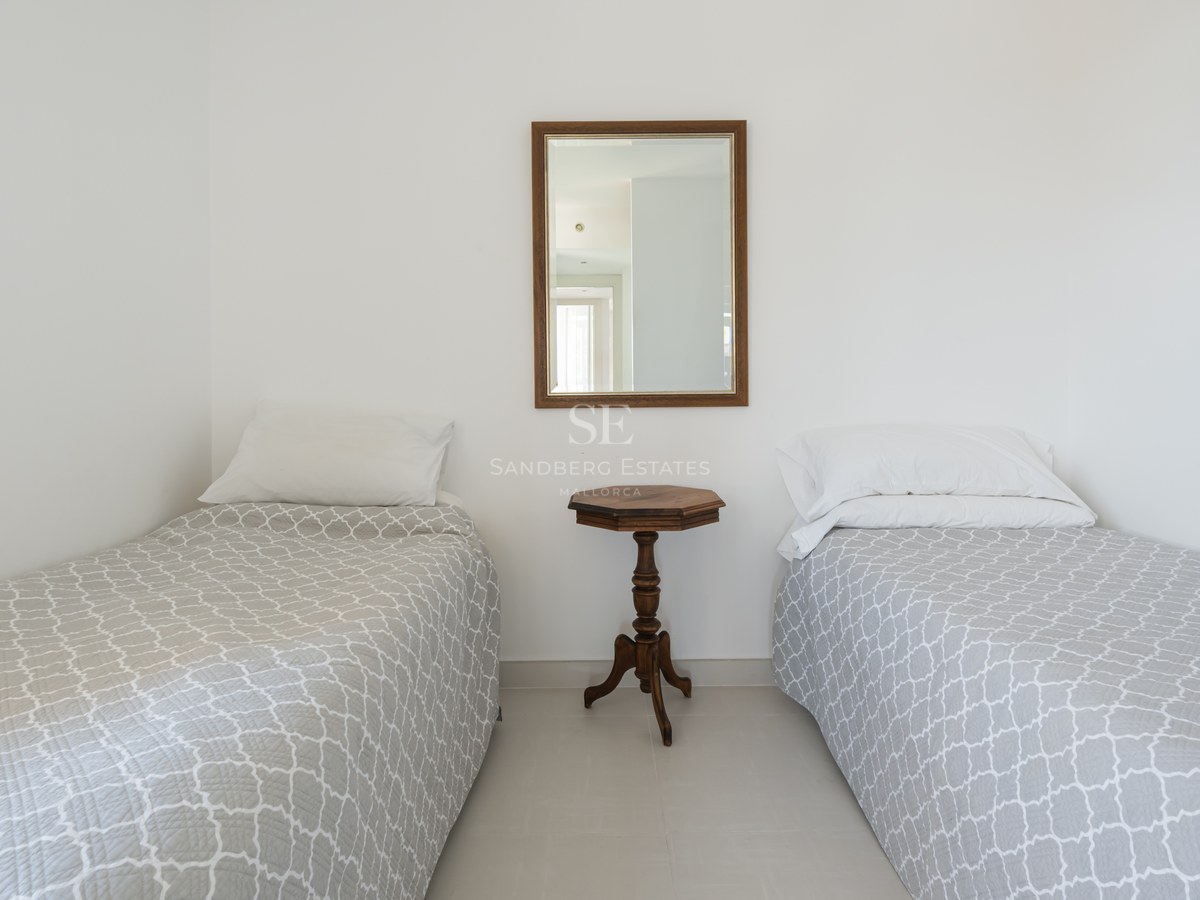 White guest bedroom featuring two twin beds with grey patterned covers and a central wooden side table and mirror.