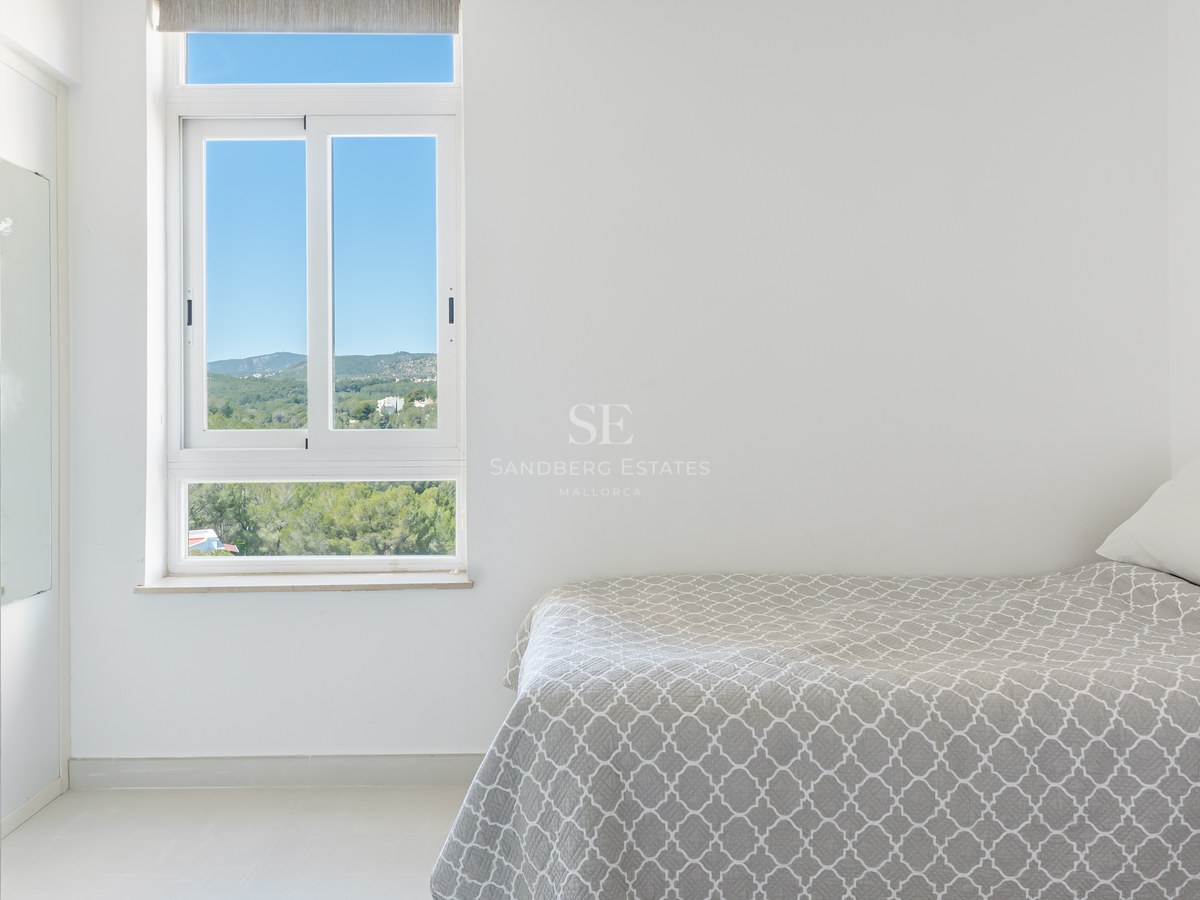 Minimalist white bedroom with a grey patterned bed and a window showing a lush green mountain landscape under a blue sky.