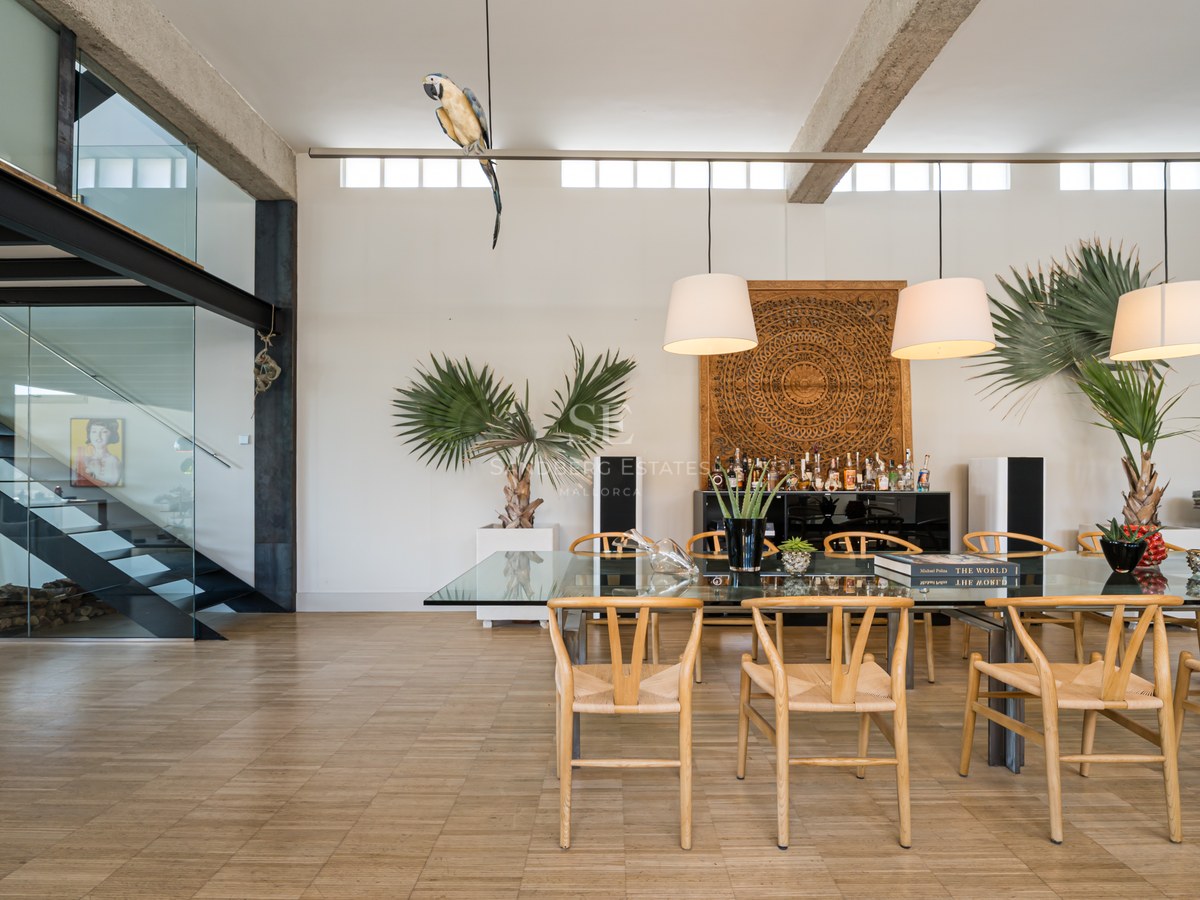 Modernes Esszimmer mit Glastisch, Holzstühlen und Stahltreppe unter hohen Decken mit Sichtbetonbalken.