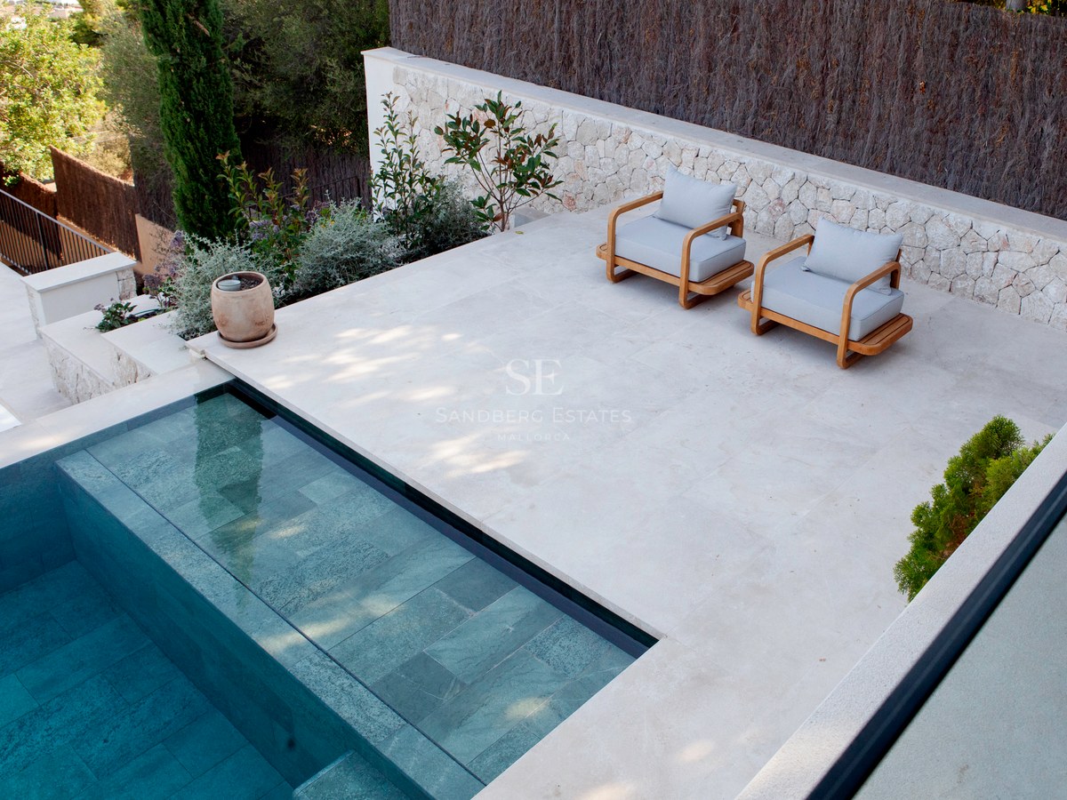 Erhöhte Ansicht eines türkis gefliesten Pools neben einer hellen Steinterrasse mit zwei Designer-Holzsitzsesseln.