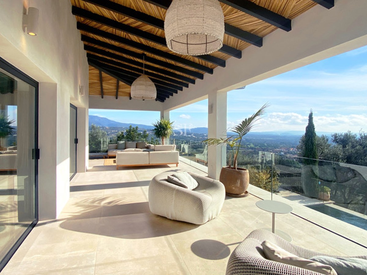 Überdachte moderne Terrasse mit beigen Loungemöbeln, Glasgeländern und weitem Blick auf ein grünes Tal und Berge.